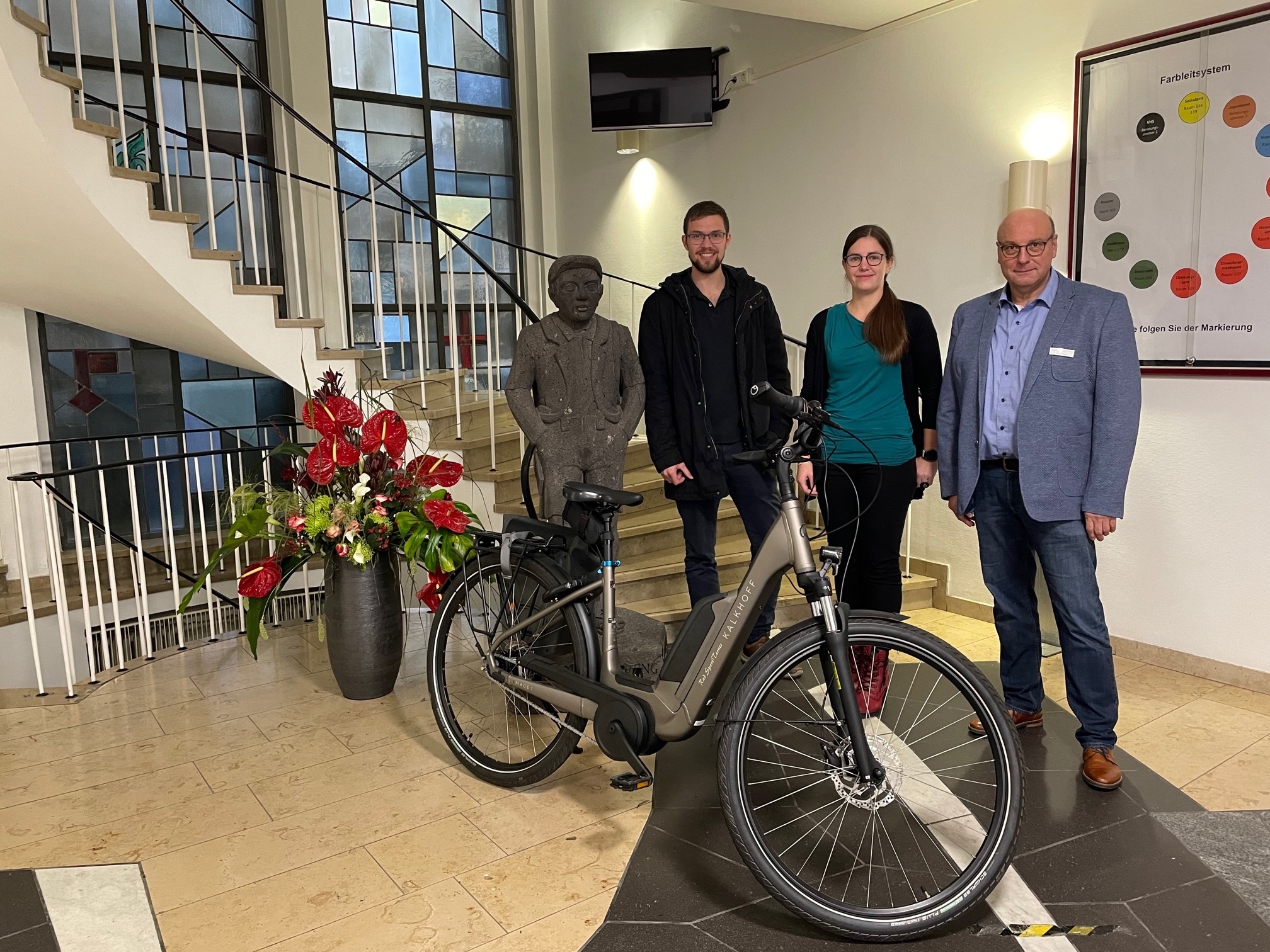 Mitarbeiter der Stadt Mayen freuen sich über Fahrrad-Leasing Zwei Männer und eine Frau stehen im Eingangsbereich der Verwaltung. Vor Ihnen steht ein E-Bike.