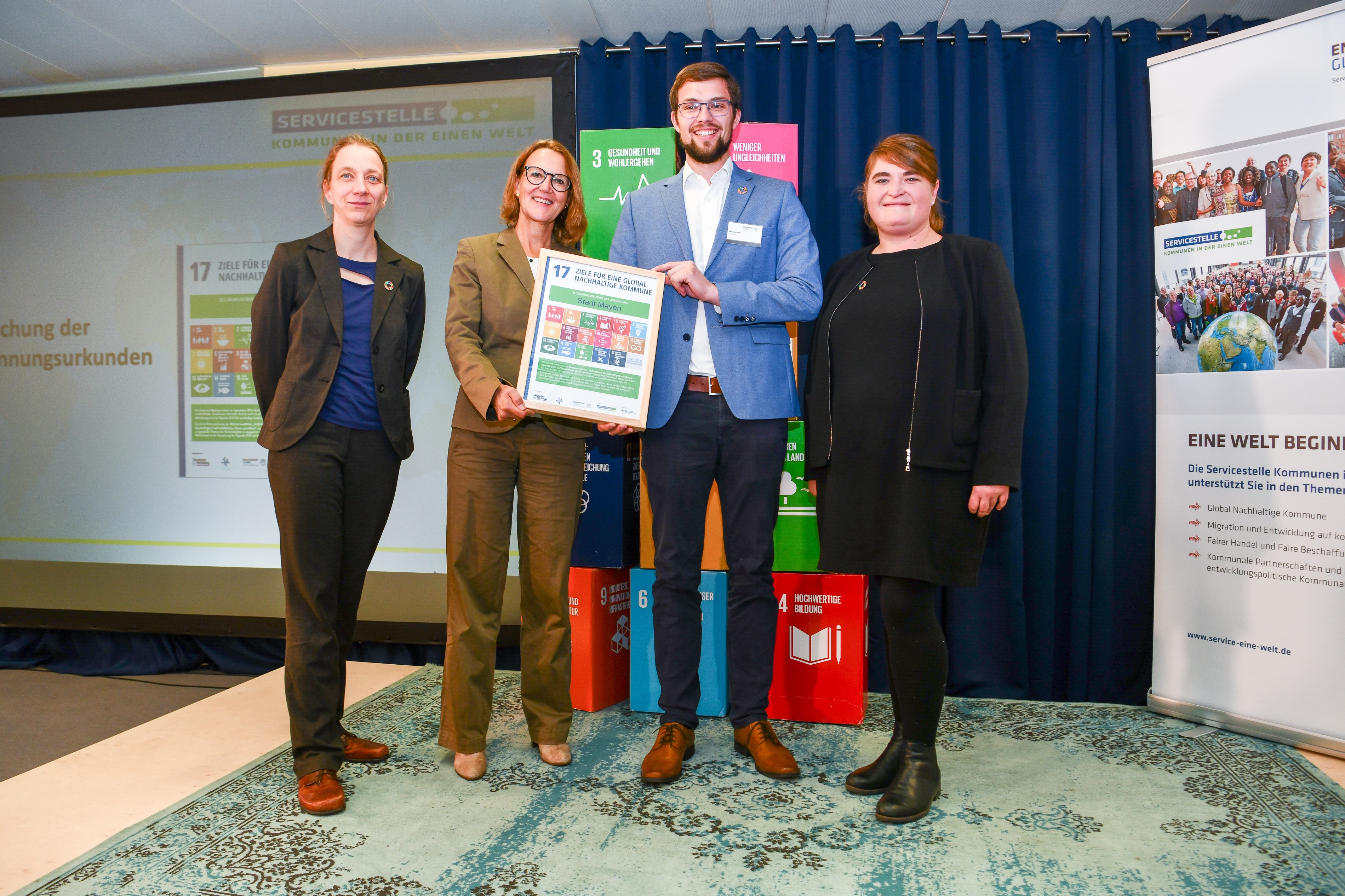 Vier Personen vor Werbebannern - Zwei davon halten eine Urkunde in der Hand