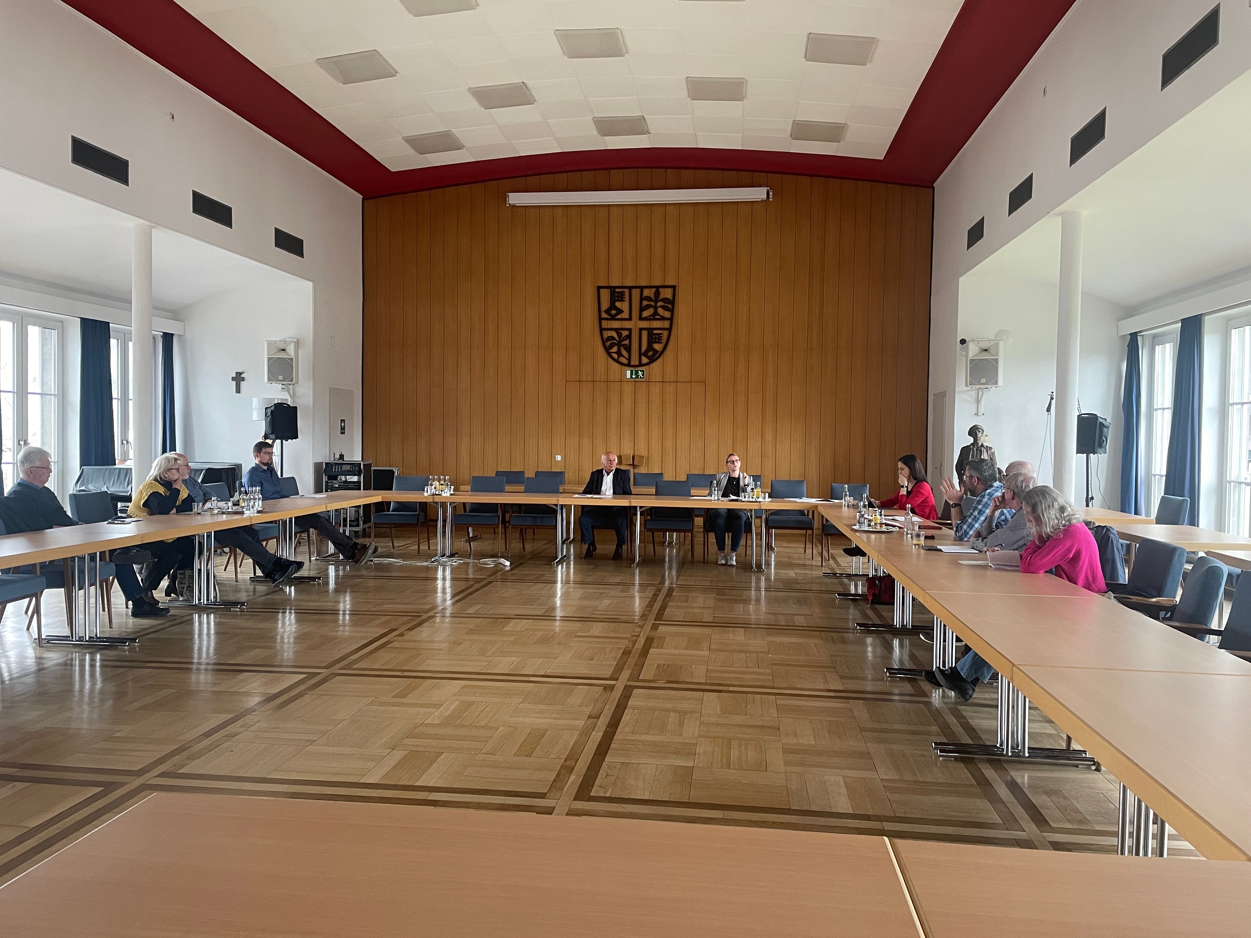 Personen sitzen an Tischen (gestellt in U-Form) zusammen und beraten sich