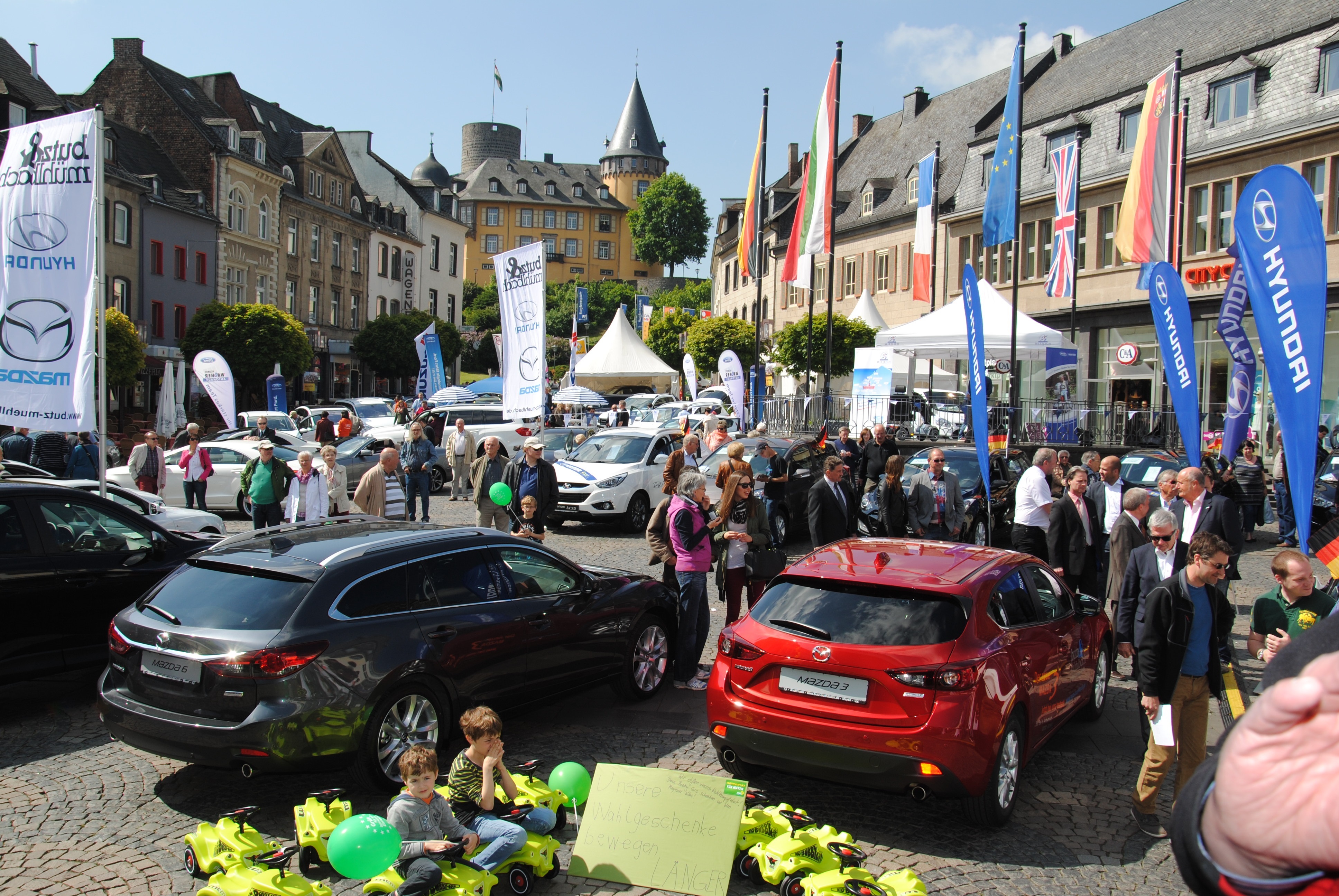 Marktplatz voller Menschen, Autos und Fahnen bei schönem Wetter