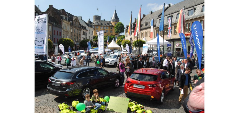 Marktplatz voller Menschen, Autos und Fahnen bei schönem Wetter