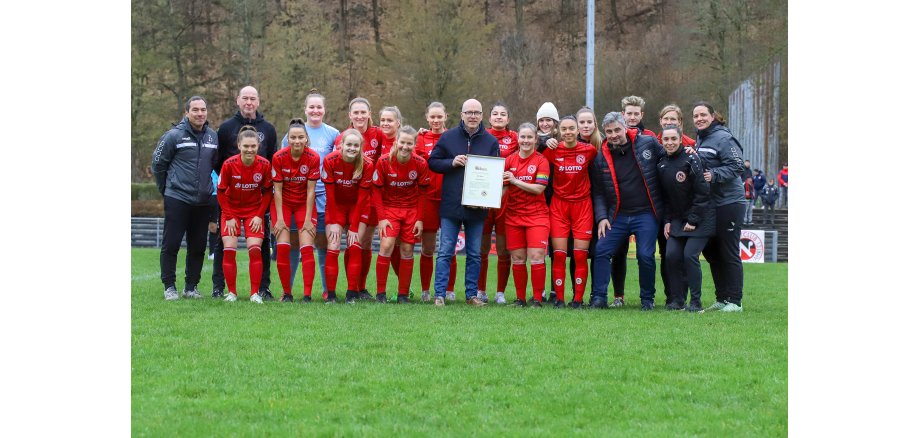 Eine Fußballmannschaft und andere Verantwortliche stehen für ein Gruppenbild auf einem Rosenplatz zusammen. Der Oberbürgermeister hält eine Urkunde in der Hand