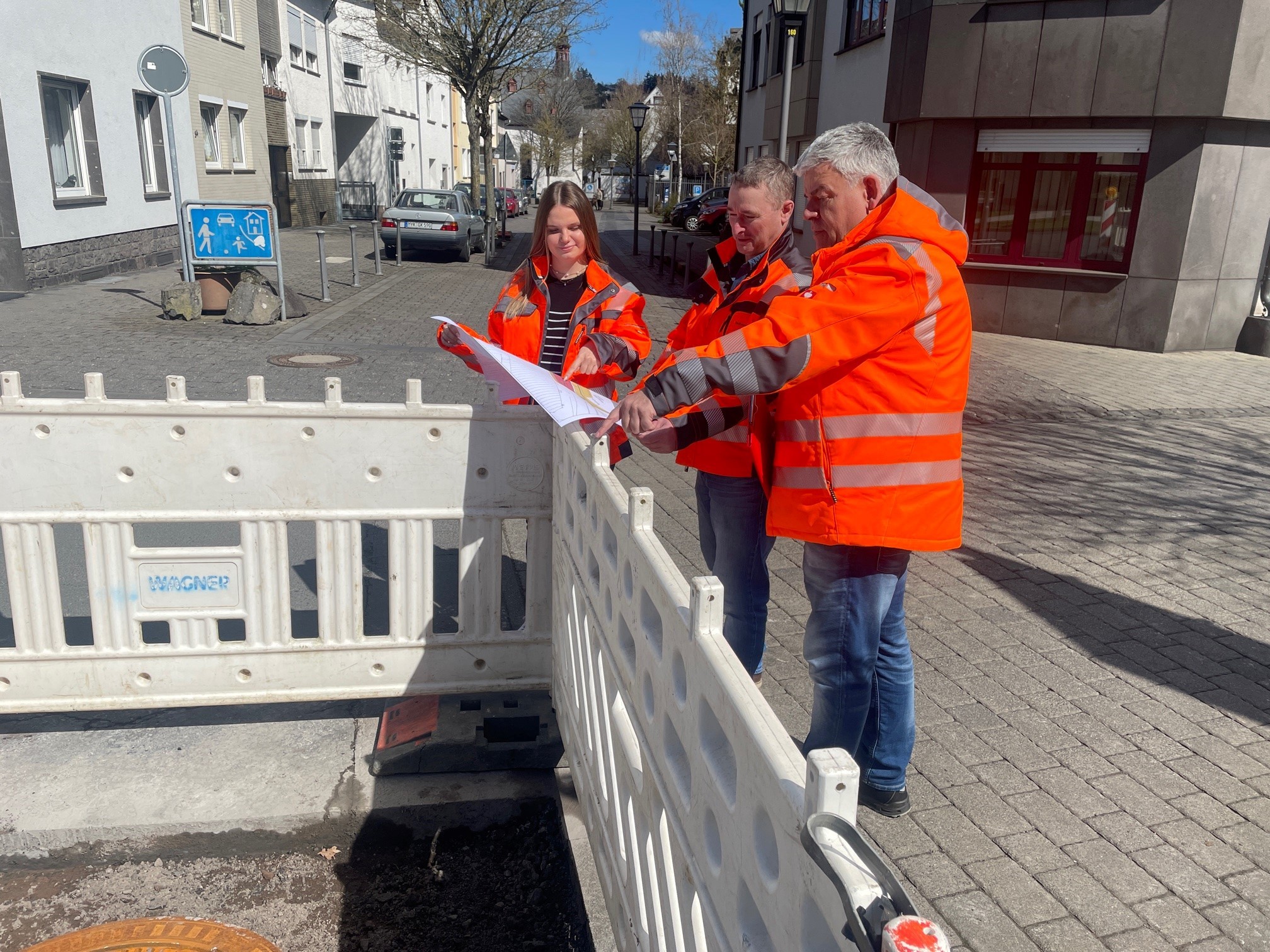 3 Personen stehen mit Sicherheitskleidung an einer Absperrung mit einem Plan in der Hand