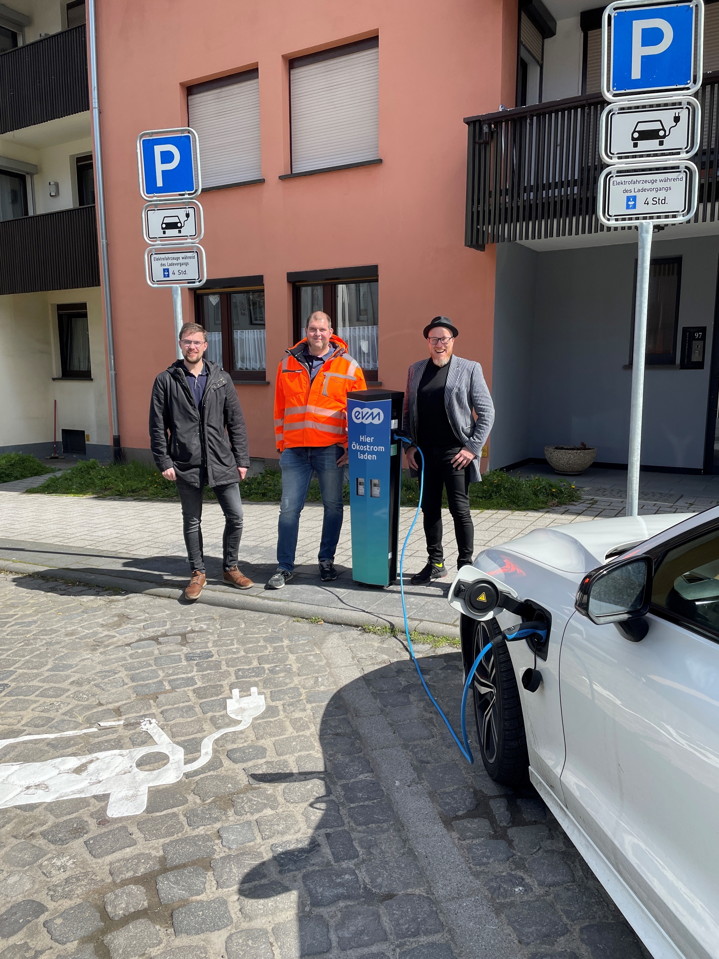 3 Personen stehen an einer E-Ladesäule vor einem Parkplatz
