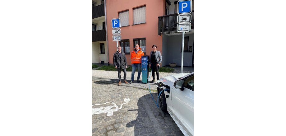 3 Personen stehen an einer E-Ladesäule vor einem Parkplatz