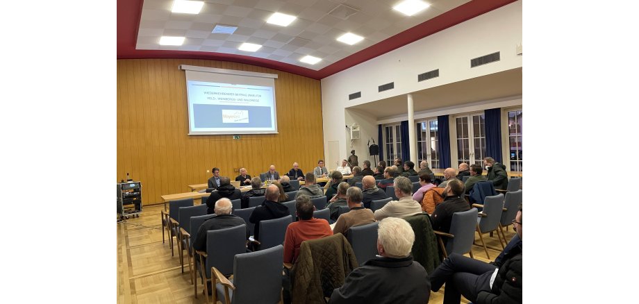 Im Sitzungssaal des Rathauses hören die Bürger zu und schauen auf einen Bildschirm mit einer Power Point Präsentation 