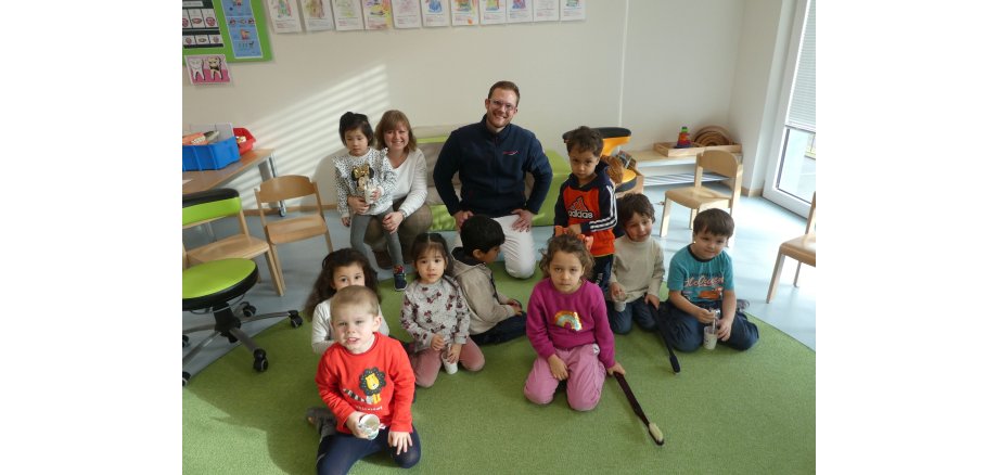 Zahnarzt Dr. Apeldorn klärt die Kinder der KiTa „In der Weiersbach“ über Zahnhygiene auf. 2 Erwachsene mit Kindern kniende auf einem Teppich