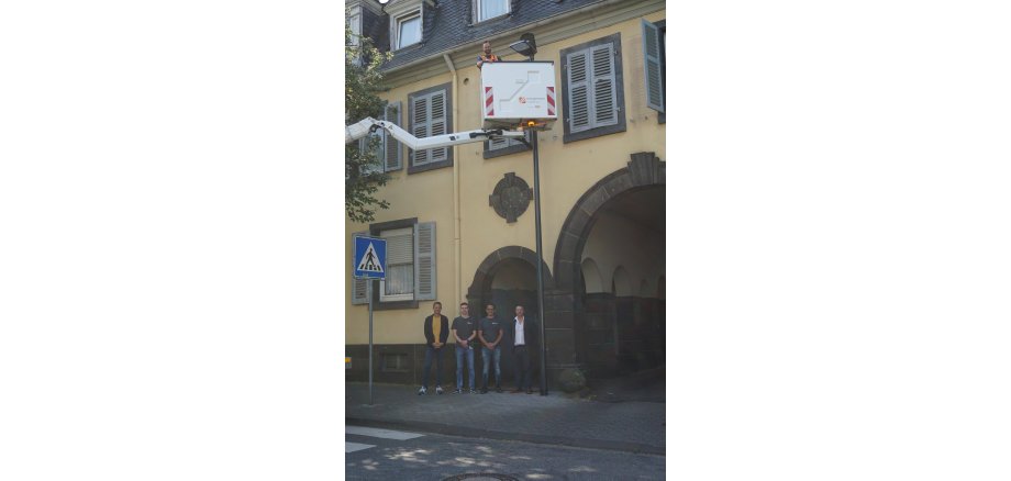 4 Männer stehen zusammen - darüber eine Person in einem Korb von einem Hubwagen an einer Laterne