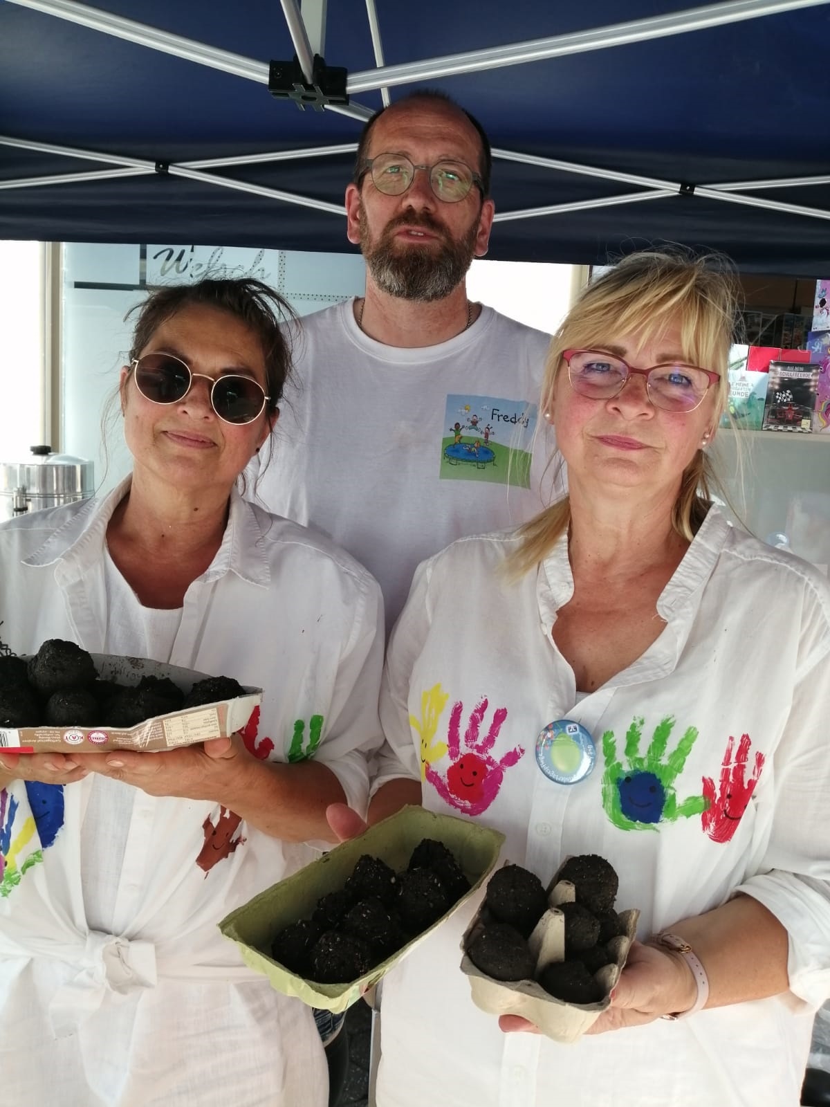 2 Frauen und ein Mann in weißen Kleidern, die bemalt sind und Kugeln aus Erde