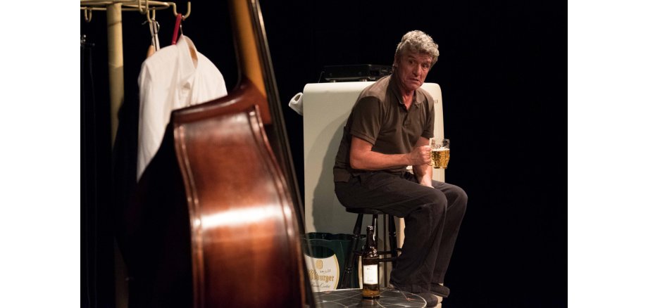 Ein Mann sitzt mit einem Glas Bier auf einem Hocker und schaut auf einen Kontrabass