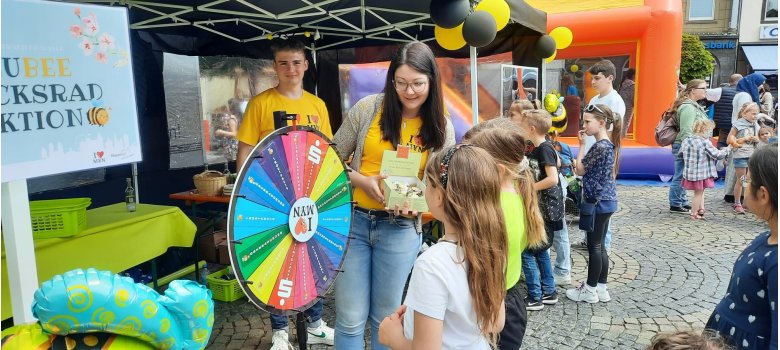 Erfolgreiche Azubiaktion bringt Spendengelder ein Einen erwachsene Person steht an einem Glücksrad mit einer Kiste Süßigkeiten in der Hand und erklärt Kindern etwas