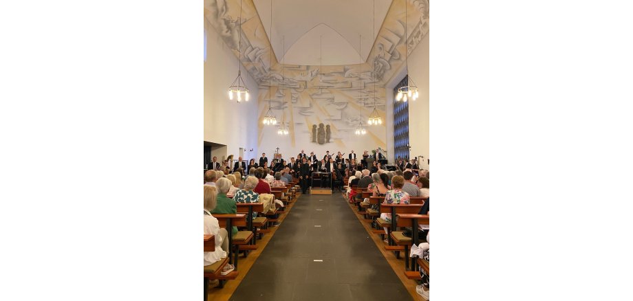 eine Gruppe von Menschen mit Instrumenten stehen vorne - davor sitzen Zuschauer auf Bänken