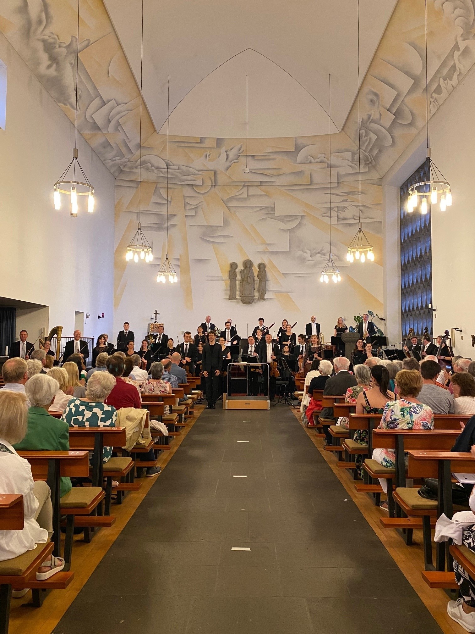 eine Gruppe von Menschen mit Instrumenten stehen vorne - davor sitzen Zuschauer auf Bänken