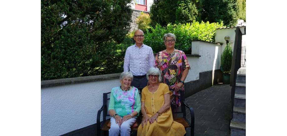 2 Frauen sitzen auf einer Bank, dahinter steht eine Frau und ein Mann für ein Foto