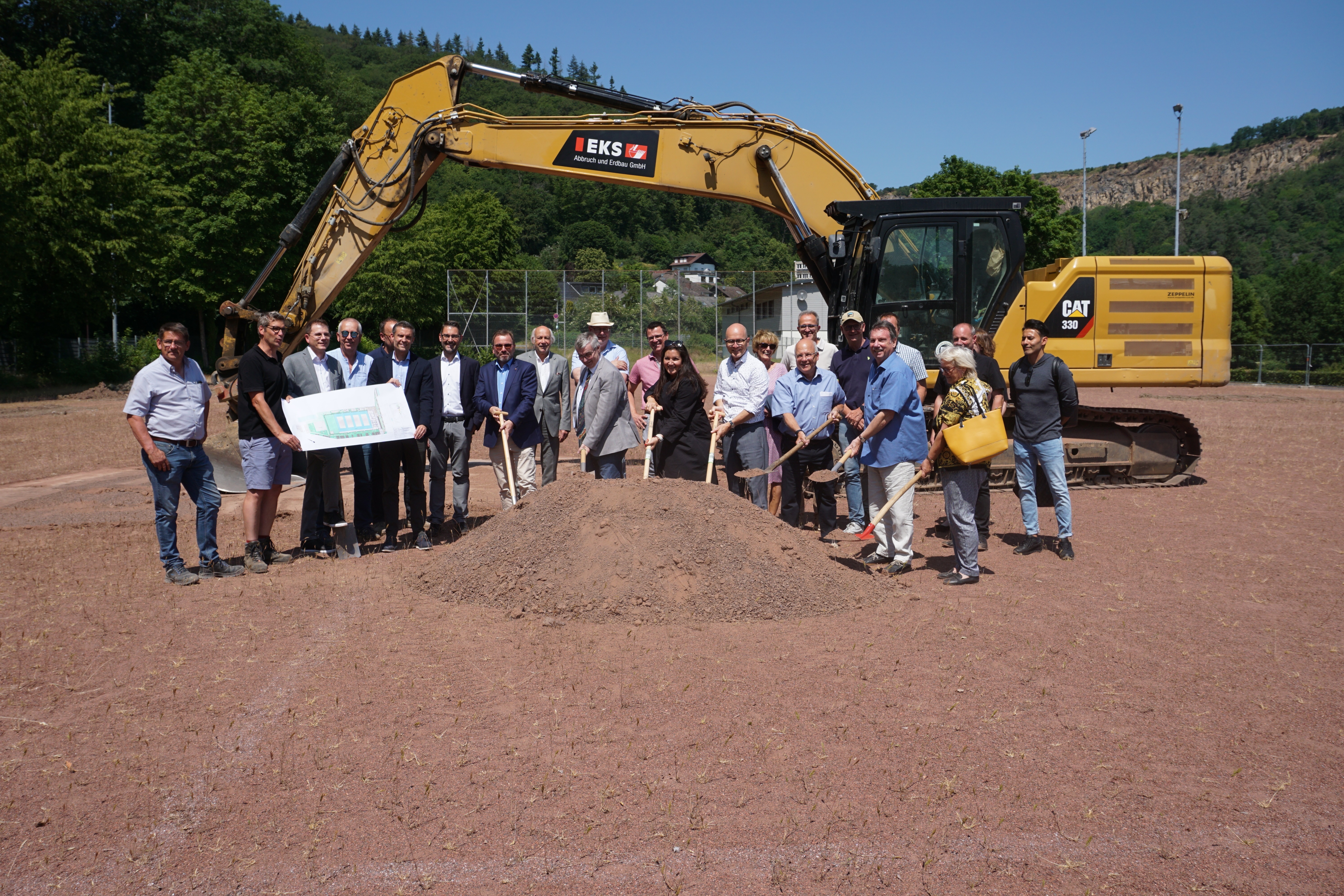 Mehrere Menschen stehen mit einer Schaufel vor einem Erdhaufen - dahinter ein Bagger