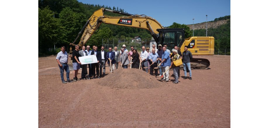 Mehrere Menschen stehen mit einer Schaufel vor einem Erdhaufen - dahinter ein Bagger