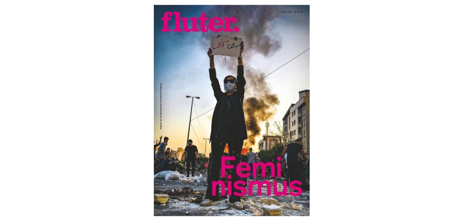 Cover des Magazins fluter Eine Frau mit Mundschutz steht inmitten von Schutt und hält ein Plakat mit arabischer Schrift nach oben. Um sie herum mehrere andere Personen und im Hintergrund ein Feuer und Rauschschwaden.