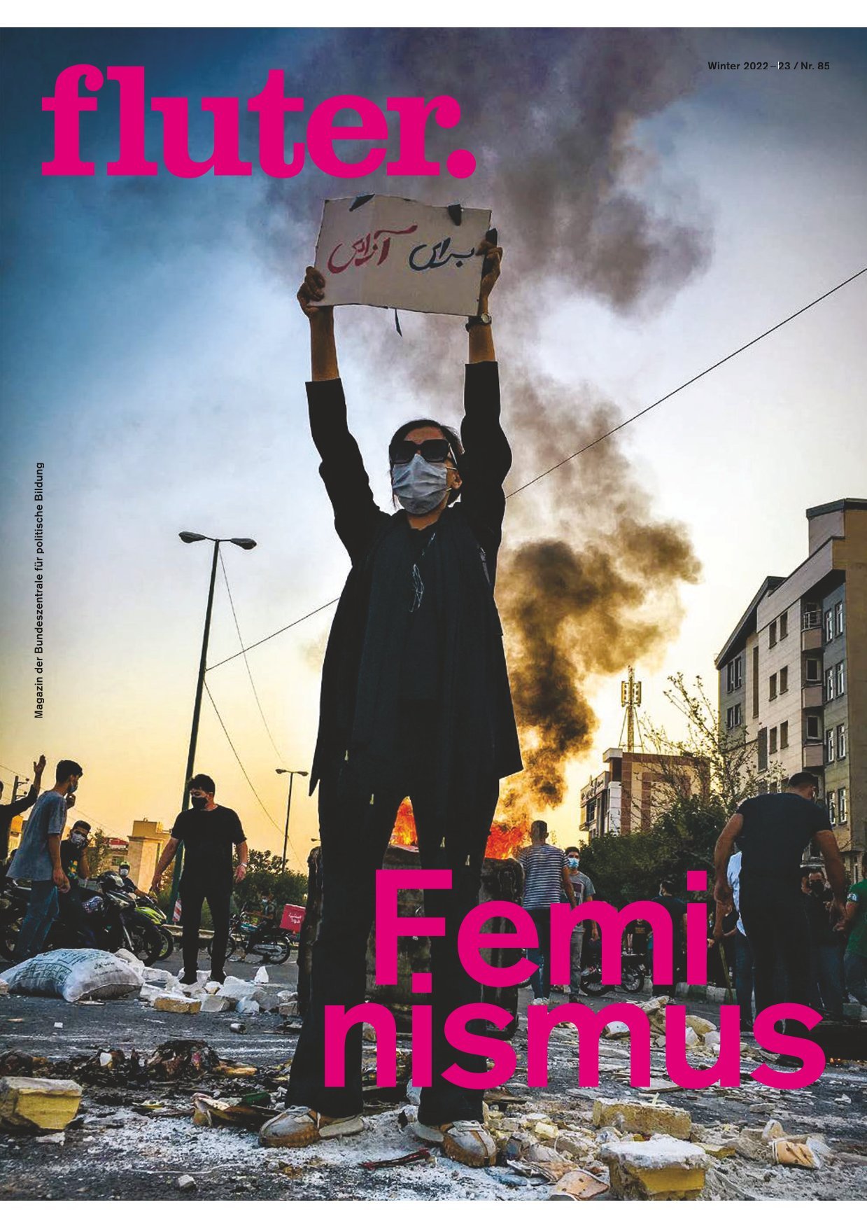 Eine Frau mit Mundschutz steht inmitten von Schutt und hält ein Plakat mit arabischer Schrift nach oben. Um sie herum mehrere andere Personen und im Hintergrund ein Feuer und Rauschschwaden. 