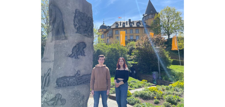 2 Personen stehen an einer Steinsäule vor einem Garten mit der Genovevaburg im Hintergrund