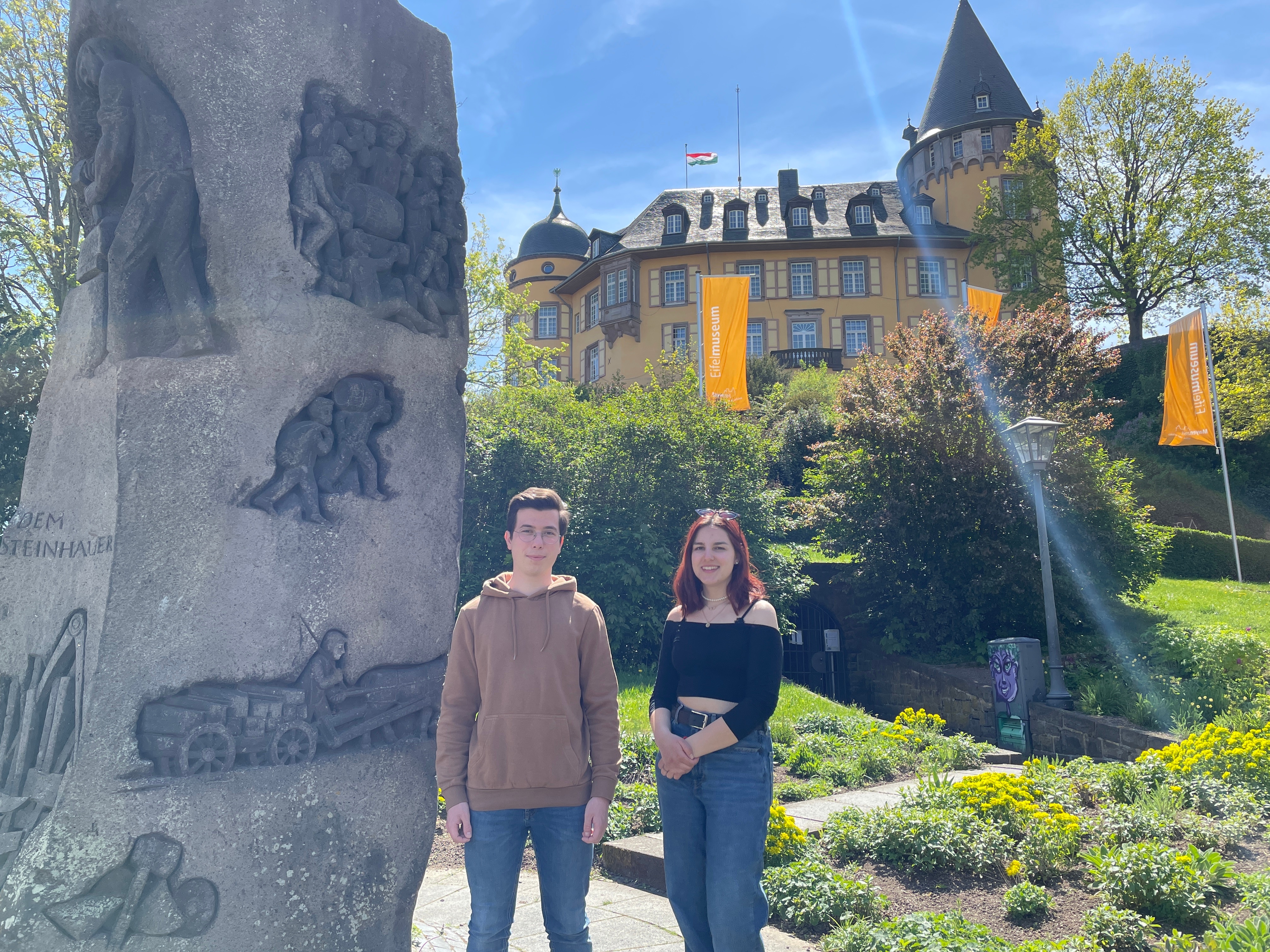 2 Personen stehen an einer Steinsäule vor einem Garten mit der Genovevaburg im Hintergrund