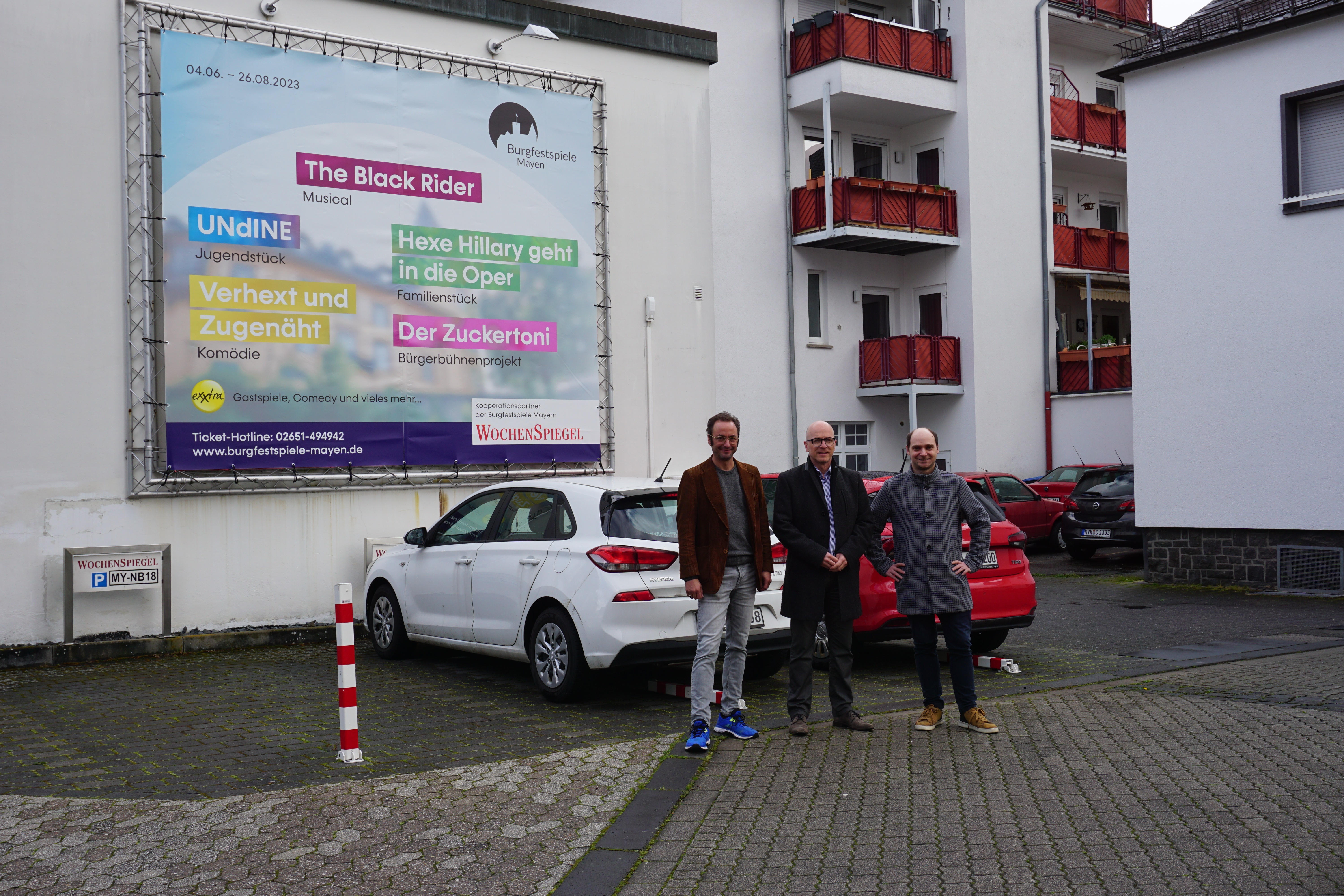 3 Personen stehe vor 2 Autos und einem Werbebanner