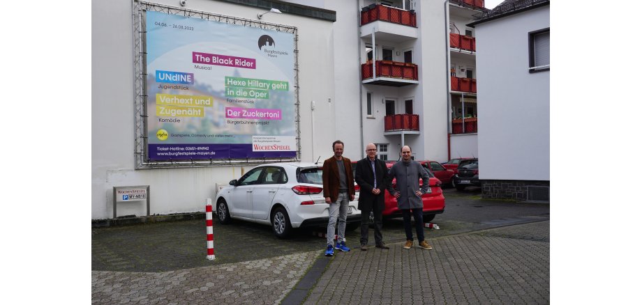 3 Personen stehe vor 2 Autos und einem Werbebanner