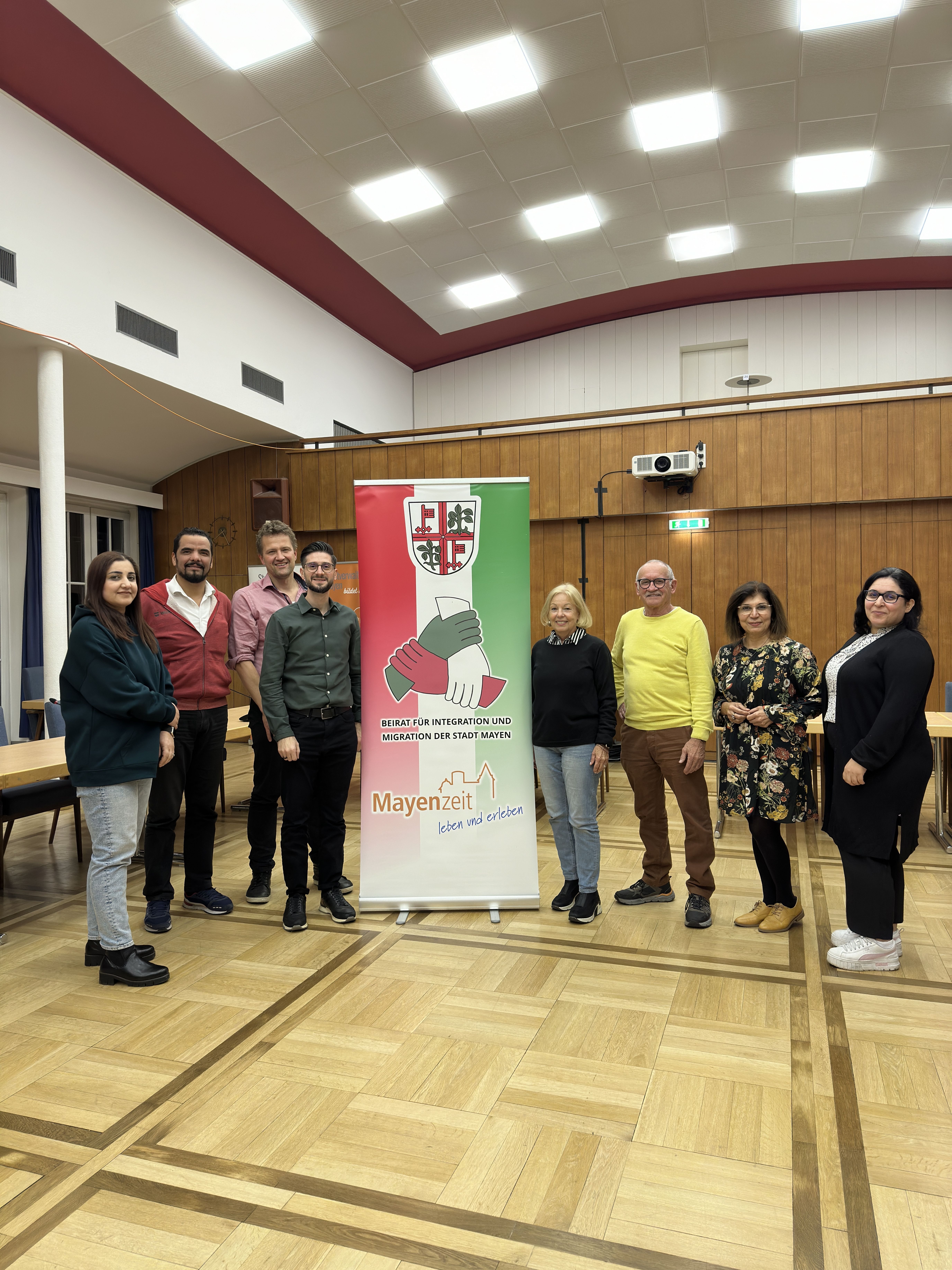 Acht Personen im Sitzungssaal neben Roll-Up des Migrationsbeirates