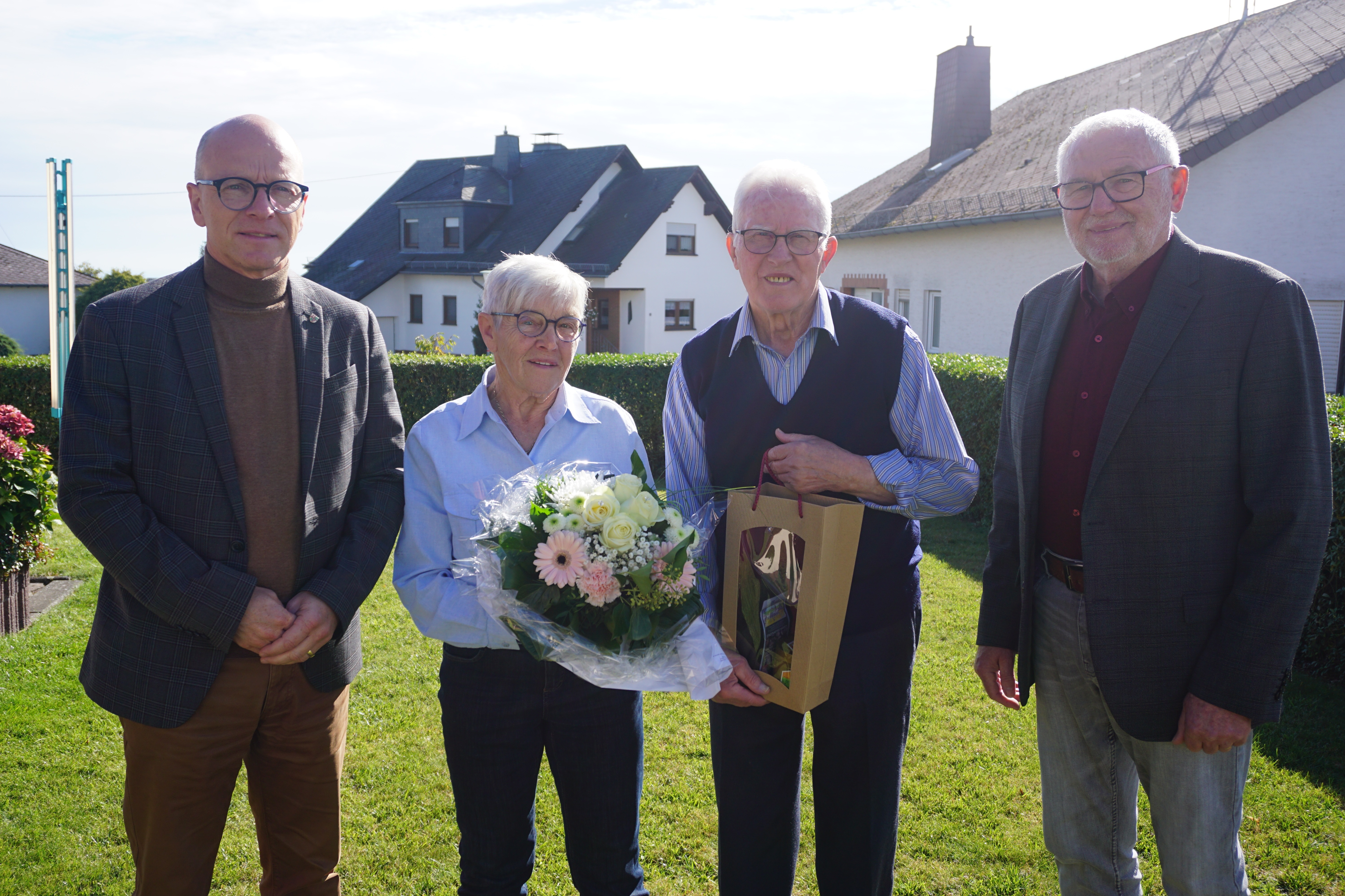 4 Personen stehen in einem Bild zusammen - die Frau hält einen Blumenstrauß in den Händen, der Mann eine Flasche Wein