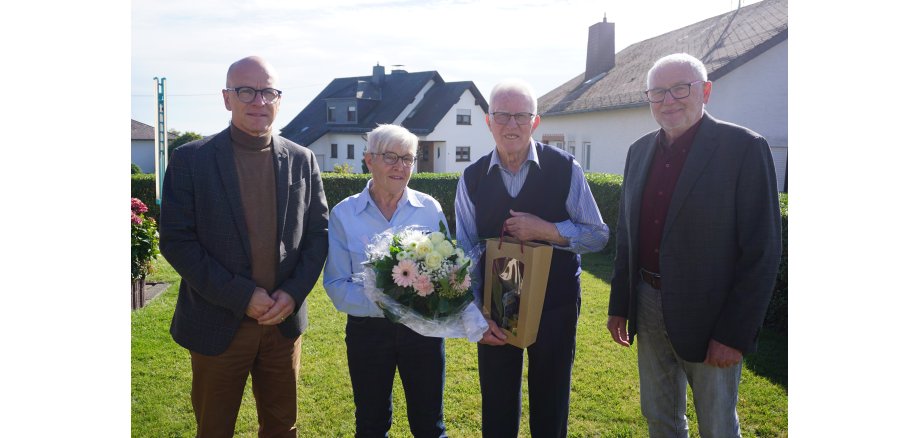 4 Personen stehen in einem Bild zusammen - die Frau hält einen Blumenstrauß in den Händen, der Mann eine Flasche Wein