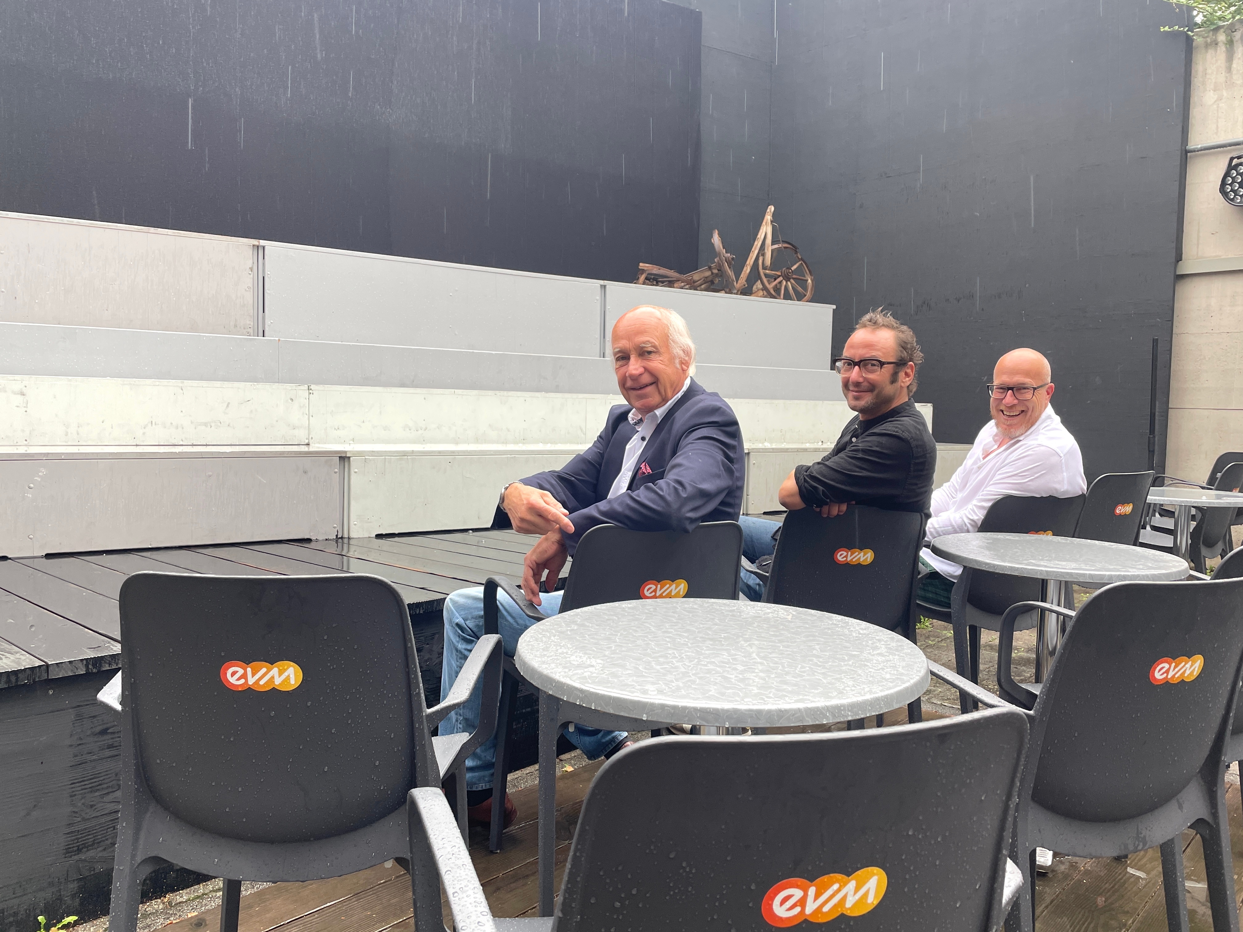 3 Personen sitzen bei Regen auf Stühlen und schauen in die Kamera