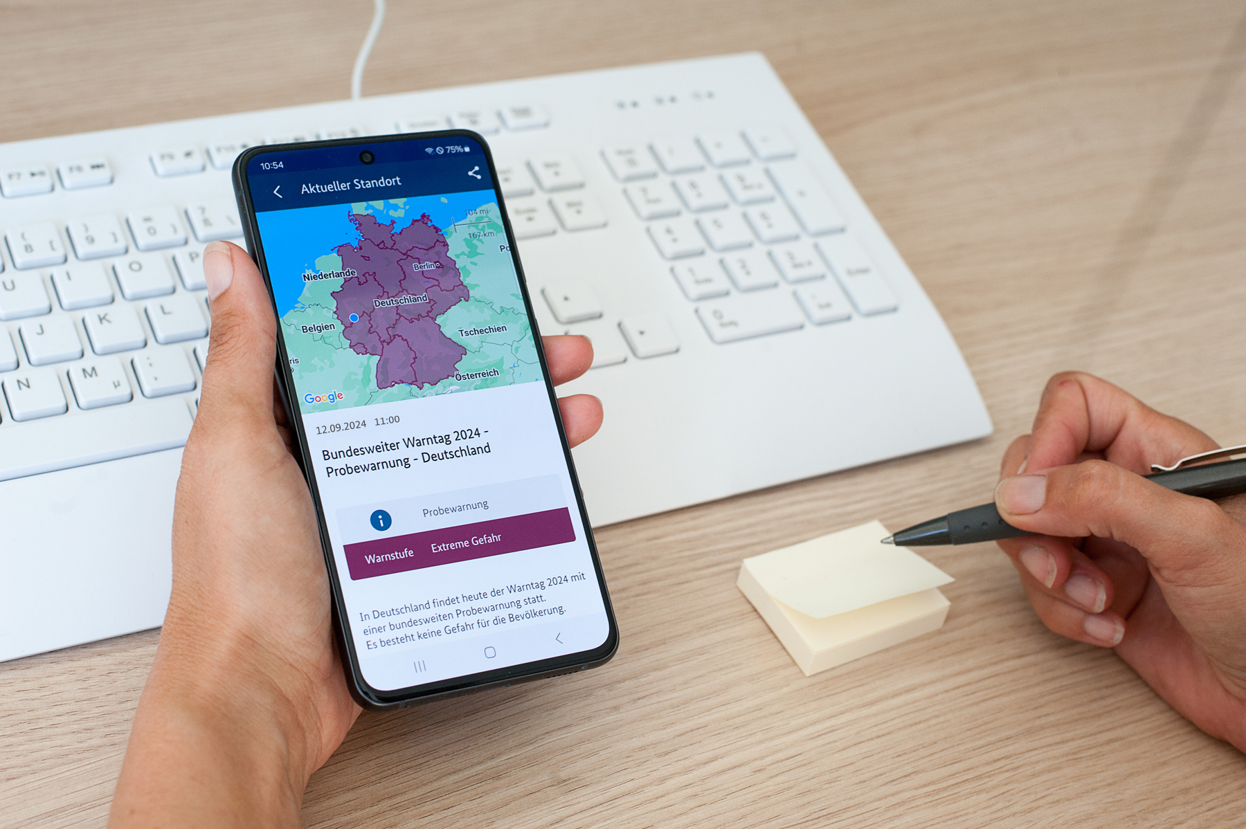 Eine Hand hält ein Handy mit einer Warnapp drauf, eine andere Hand hält einen Kugelschreiber
