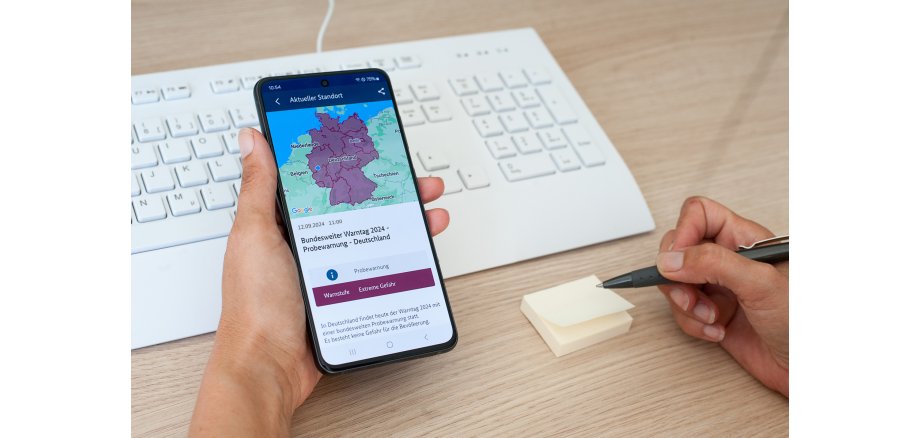 Eine Hand hält ein Handy mit einer Warnapp drauf, eine andere Hand hält einen Kugelschreiber