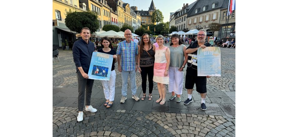 7 Personen stehen zusammen - 2 davon halten ein Plakat der Summer Beats in den Händen