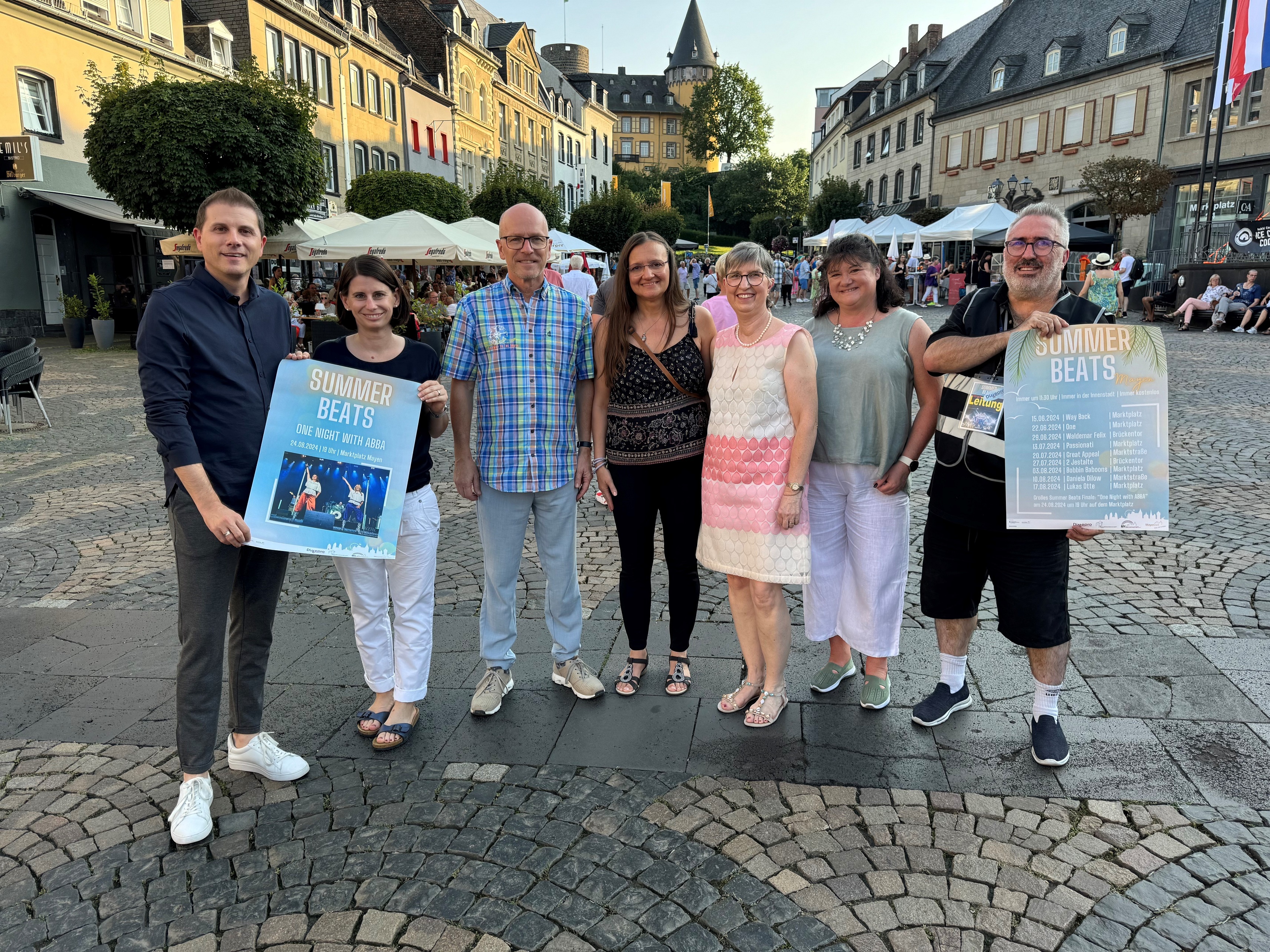 7 Personen stehen zusammen - 2 davon halten ein Plakat der Summer Beats in den Händen