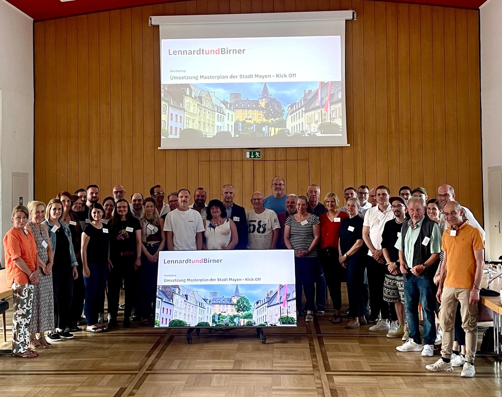 Gruppe von Menschen steht im Sitzungssaal vor einer Leinwand und einem großen Bildschirm