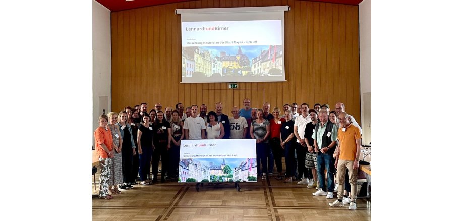 Gruppe von Menschen steht im Sitzungssaal vor einer Leinwand und einem großen Bildschirm