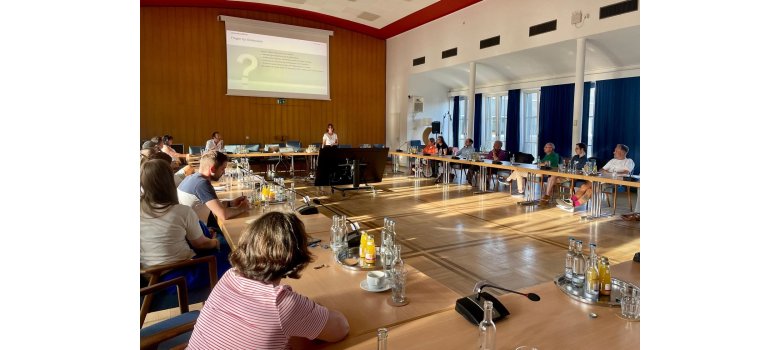 Menschen sitzen an Tischen und schauen auf eine Leinwand - vor der Leinwand steht eine Frau