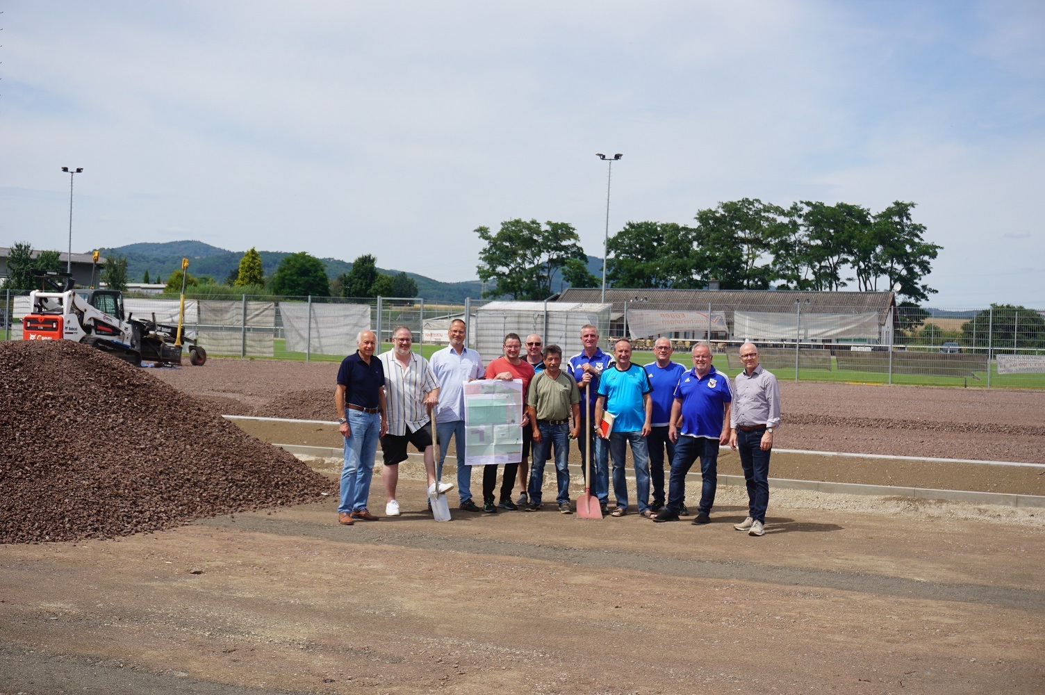 Mehrere Personen stehen auf einem Platz neben einem Schotterhaufen - einige haben ein Plakat und Schaufeln in der Hand