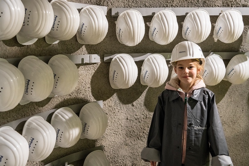Ein kleines Mädchen hat einen Sicherheitshelm auf dem Kopf und steht vor einer Wand mit Helmen