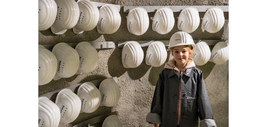 Ein kleines Mädchen hat einen Sicherheitshelm auf dem Kopf und steht vor einer Wand mit Helmen