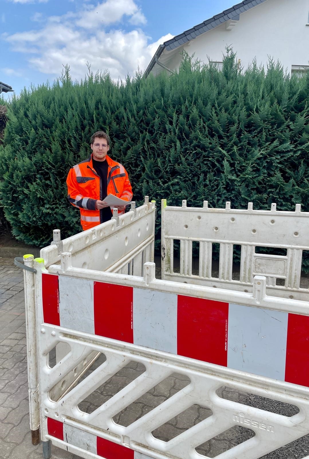Ein Mann steht mit einer gut sichtbaren Arbeitsjacke an einer Baustellenabsperrung und hat Unterlagen in der Hand