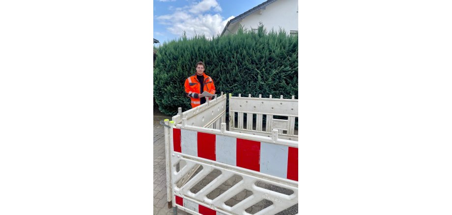 Ein Mann steht mit einer gut sichtbaren Arbeitsjacke an einer Baustellenabsperrung und hat Unterlagen in der Hand