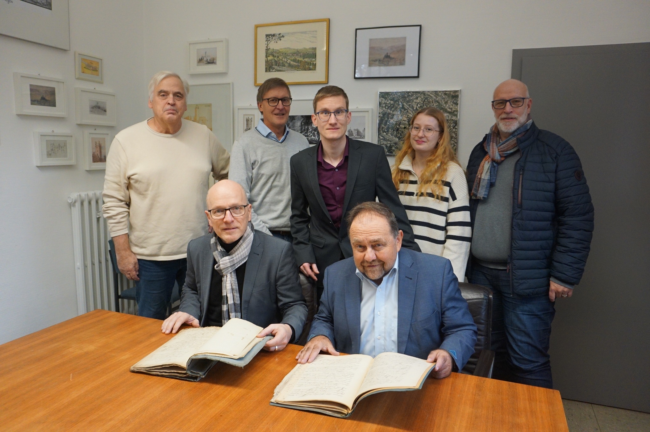 2 Personen sitzen an einem Tische eine weitere zwischen Ihnen. Dahinter stehen 4 weitere Personen