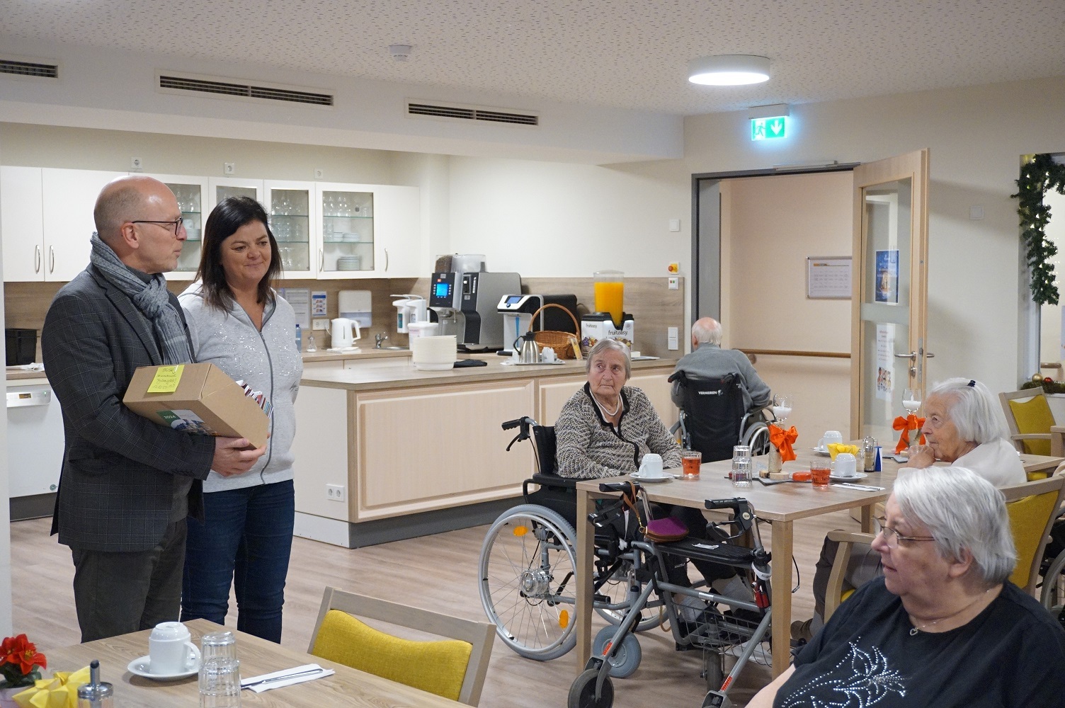 Zwei Personen stehen mit Geschenken in einem Raum - mehrere Seniorinnen und Senioren hören aufmerksam zu