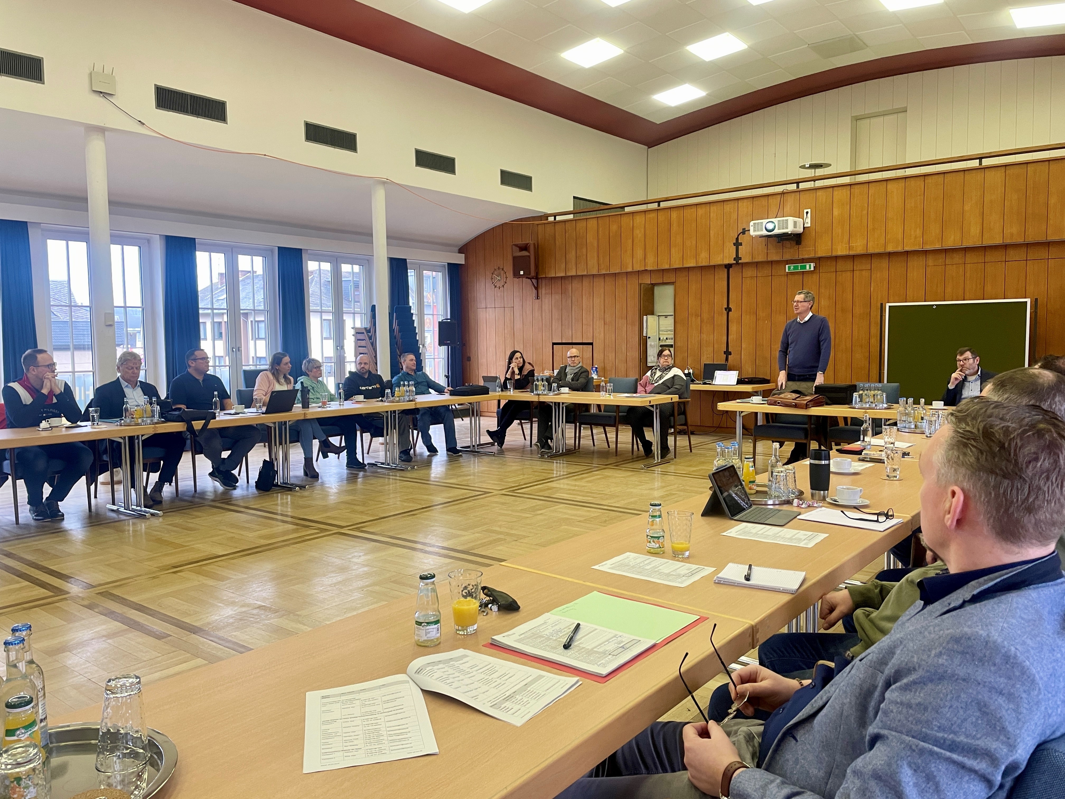 Menschen sitzen in U-Form an Tischen und hören einem Dozenten zu