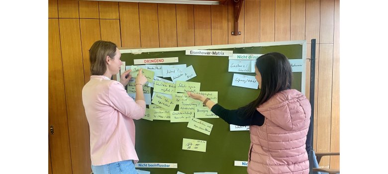 Zwei Frauen stehen vor einer Pinnwand und pinnen Notizen an die Wand