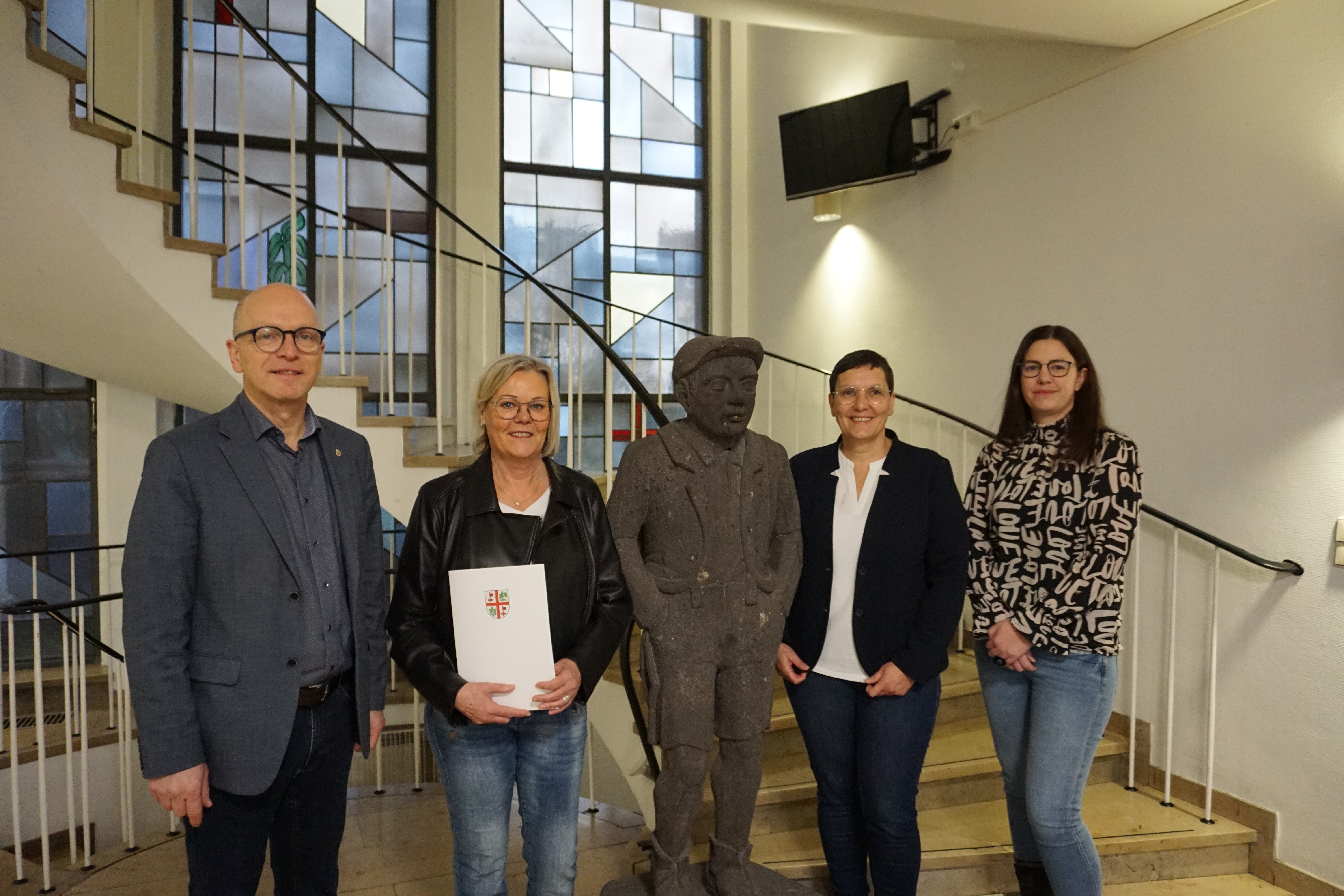 4 Personen stehen um eine Steinfigur vor einer Treppe zusammen - eine Frau hält eine Urkunde in der Hand