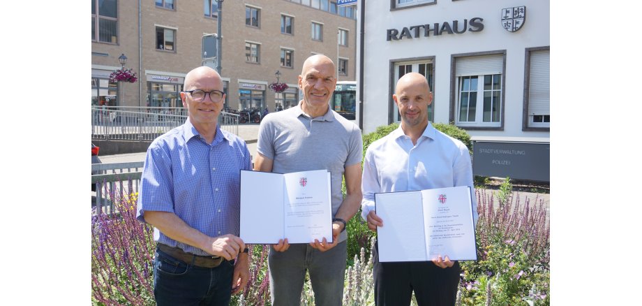 3 Männer stehe vor dem Rathaus und 2 davon halte eine Urkunde in den Händen