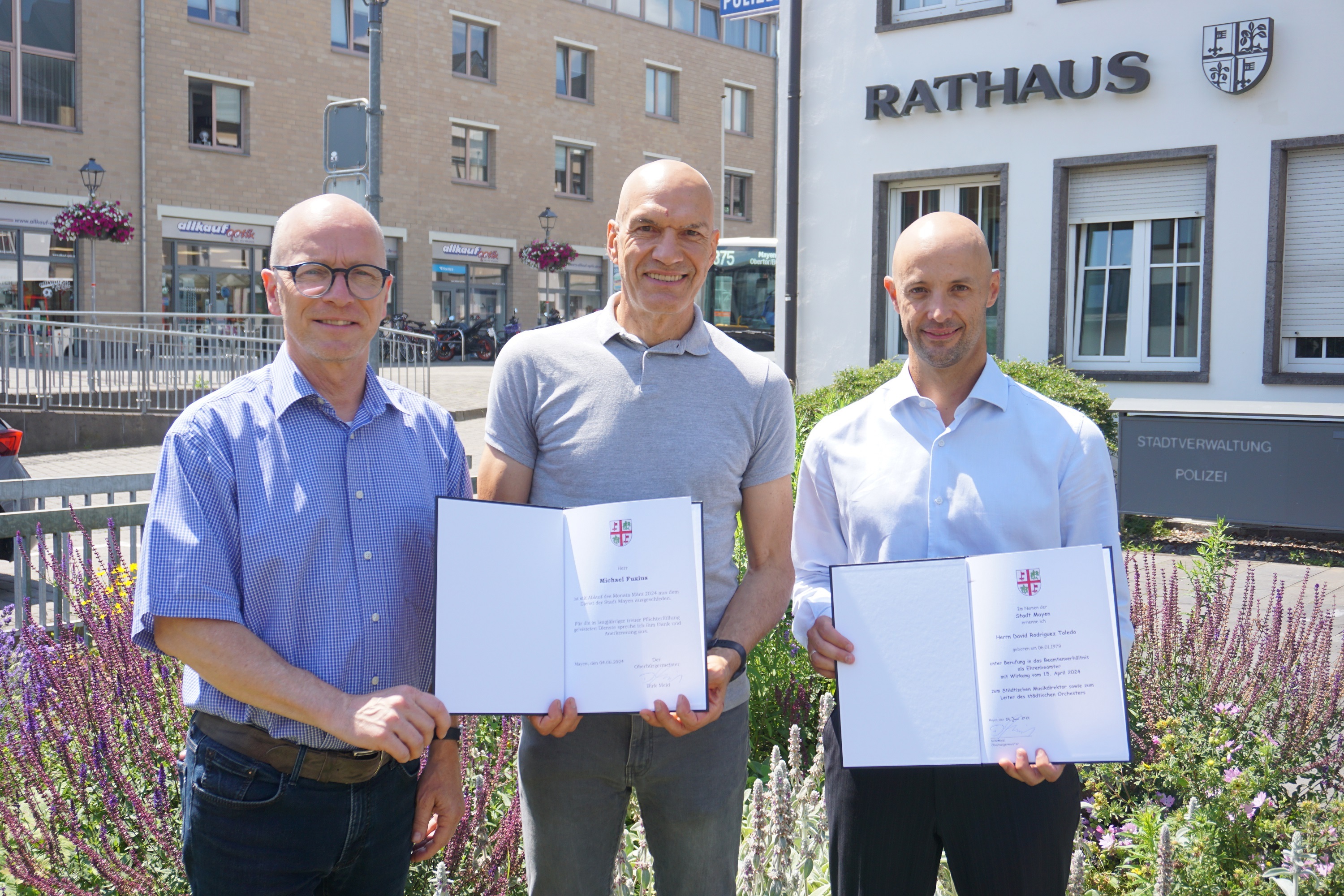 3 Männer stehe vor dem Rathaus und 2 davon halte eine Urkunde in den Händen