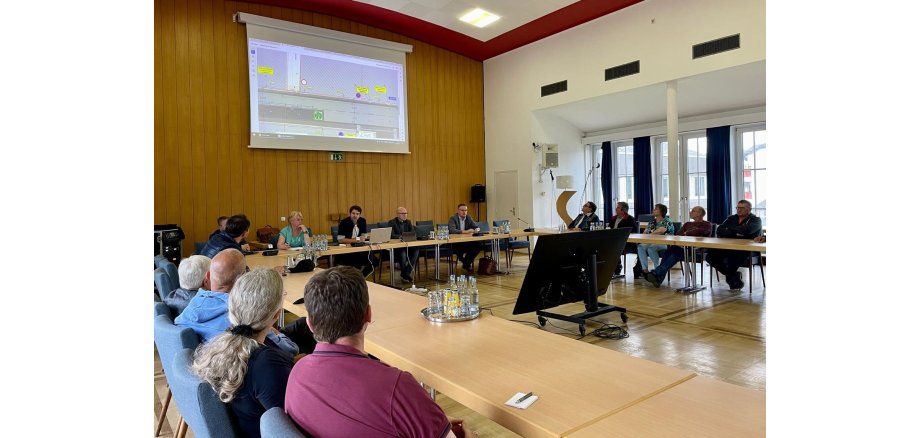 Personen sitzen an einem Tisch und verfolgen eine Präsentation auf der Leinwand